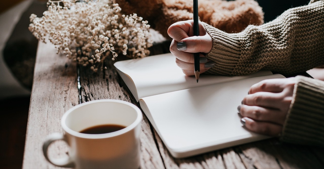 Woman writing in a journal with a cup of coffee, flowers, and teddy bear next to the journal on a table